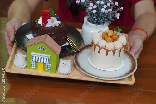 A woman holds a wooden tray containing macadamia coffee cake, chocolate cake, and a vase with pink flowers, brown wooden table