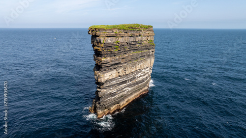Photography Aerial view of a towering sea stack in Ireland with layered rock cliffs rising from blue water of the Atlantic Ocean beneath a clear sky