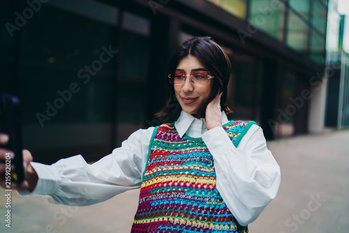 Hispanic woman in pink glasses posing for smartphone selfie photo on city street copy space. Creative latin female content creator adjusting hair, posting to social media channel network.