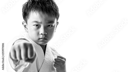Focused young Asian boy practicing a punch in a traditional white karate uniform, showing discipline and strength in this black and white portrait against a clean background. Copy space on the right.