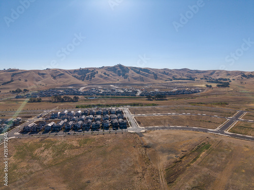 Aerial view showing a new housing development growing into a dry, arid landscape with future expansion plots and rolling hills under a clear blue sky, illustrating urban sprawl and growth