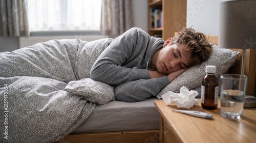Sick teenager resting in bed with tissues, thermometer and medicine nearby during cold and flu recovery.