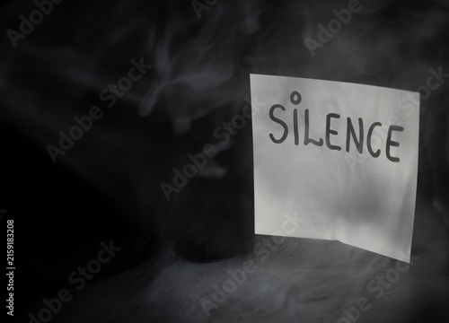 Konstfotografi White sticky note with handwritten word SILENCE in dramatic swirling smoke on dark background
