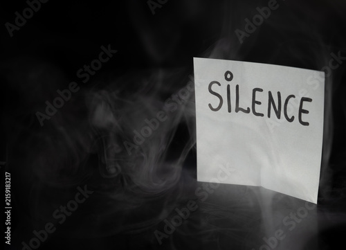 Konstfotografi White sticky note with handwritten word SILENCE in dramatic swirling smoke on dark background