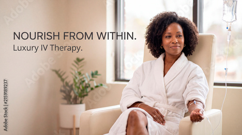Smiling Black woman in a white bathrobe sitting on an armchair receiving intravenous IV therapy drip in a bright wellness clinic, luxury health care treatment, peaceful recovery moment.