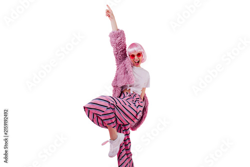 Blissful woman wears striped pants and pink periwig laughing during photoshoot. Confident young lady in sunglasses and fluffy jacket funny dancing.