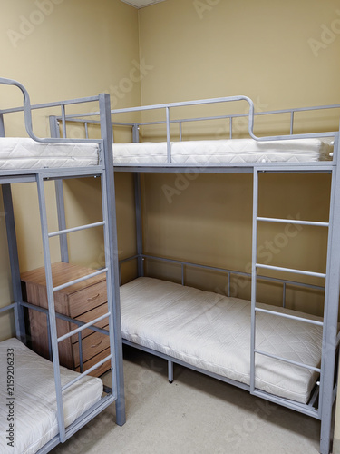 Metal bunk beds with empty mattresses in a simple room featuring beige walls and a wooden nightstand, suitable for dormitory or shared living spaces