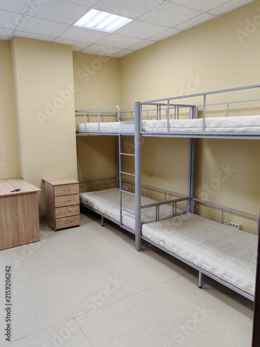 Bunk beds with mattresses in a simple room featuring beige walls, a wooden dresser, and a bright overhead light in a shared living space