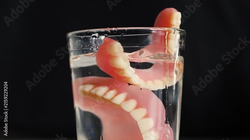 A second full denture falls into a glass of water that already contains the first one, causing splash on a black background. Demonstration of daily cleaning 