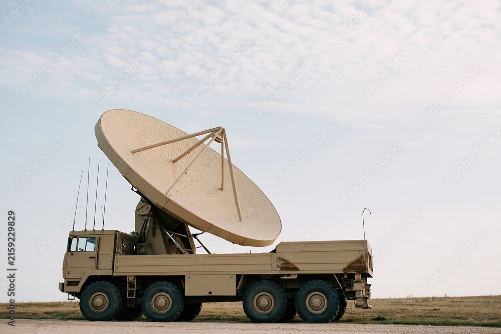 Naklejka premium Large parabolic antenna on a heavy truck chassis, featuring a weathered matte finish and textured mesh for communications infrastructure and defense technology, with copy space