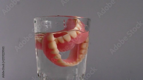 A second full denture falls into a glass of transparent water that already contains the first one, causing a splash. 