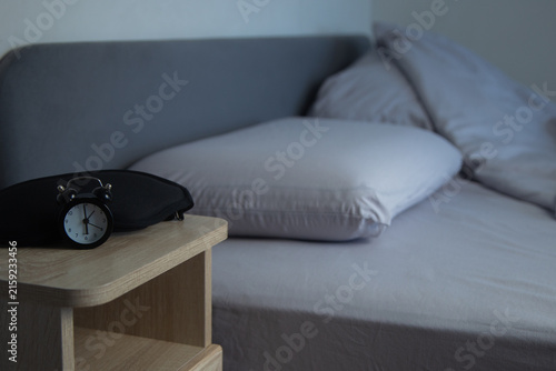 On a light wooden bedside table lies a black sleep mask with a miniature black alarm clock on the background of a bed with gray pastel linens. The concept of a sleep preparation atmosphere. High