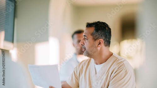 Middle Eastern male nurse in beige scrubs glancing at a wall screen, showing tired but steady competence in a hospital for healthcare and clinical documentation, copy space