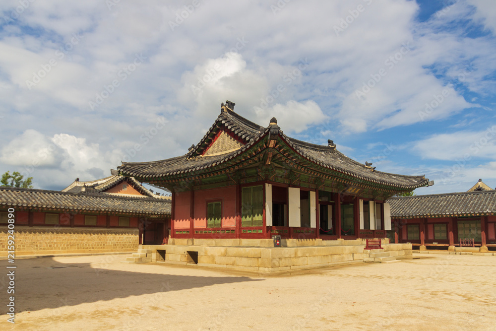 Fototapeta premium Ornate traditional buildings with tiled roofs and painted wooden eaves surround a sunlit stone courtyard beneath a cloud-filled blue sky.