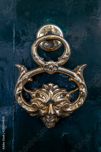 An ornate brass door knocker shaped like a horned face flanked by dolphin-like scrolls hangs against a glossy dark green door in Valletta, Malta