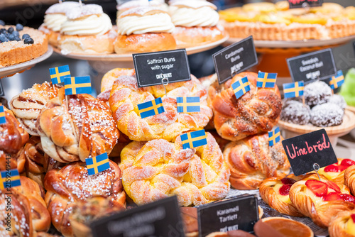 Assorted traditional Swedish pastries including saffron buns, cinnamon buns, semla and fruit pastries, decorated with Swedish flags in a colorful bakery display.