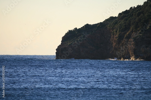Canvastavla Scenic coastal landscape featuring a steep, vegetated cliff dropping into the deep blue sea with white foam waves crashing at the base