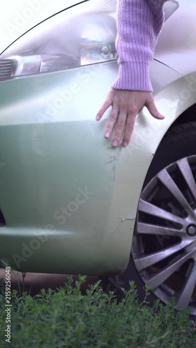Woman's hand examines scratches and cracks on a light green car bumper after an accident. Outdoor close-up showing wheel and bushes.