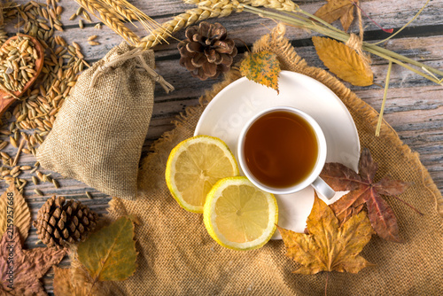Warm autumn herbal tea with lemon slices, dry leaves, pine cones, grains, and burlap sack on rustic wooden background top view flat lay