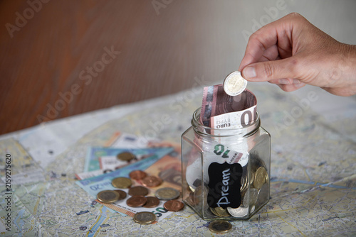 Travel and financial planning concept with a miniature globe, glass jar filled with euro banknotes and coins, and maps, symbolizing tourism, vacation budgeting, international currency, savings and tra