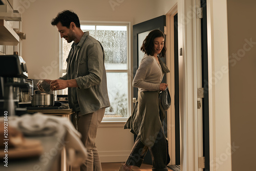 Man prepares breakfast in sunlit home kitchen while woman enters doorway, showing calm morning routine, casual clothing, warm interior light, modern apartment and everyday domestic comfort