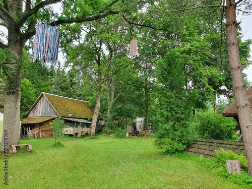 A rustic rural scene featuring a traditional wooden barn with a moss-covered roof situated in a lush green clearing surrounded by dense forest. Colorful woven ribbons hang from tree branches as