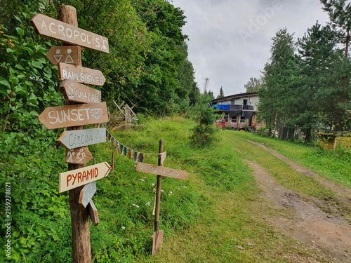 A rustic wooden signpost with multiple directional arrows stands beside a dirt path in a lush green rural setting. The signs point to various whimsical destinations including 