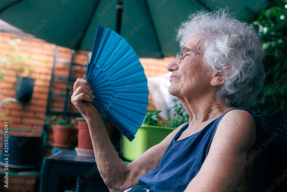 Fototapeta premium Elderly woman cooling herself with a fan while sitting on the balcony during a heatwave