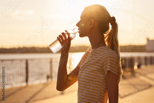 Cuadro en lienzo Young woman drinking water from a bottle during sunset, hydration and healthy li