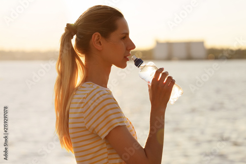 Fotografía Young woman drinking water from a plastic bottle outdoors during golden hour, hy