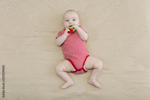 Baby lying on bed and gripping teething ring with both hands. Child chewing toy for teething relief. Exploring textures while soothing sore gums. Sensory discovery at home.