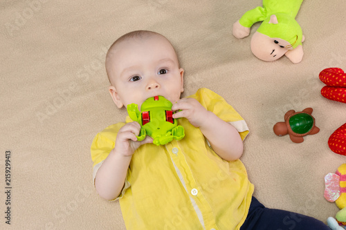 Cute 9 month baby boy lying among various playful colorful toys on beige blanket. Infant surrounded by vivid objects participating actively in sensory exploration and learning. 