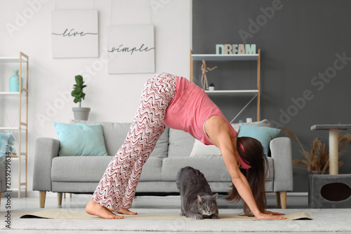 Young pregnant woman with cute cat doing yoga on mat at home