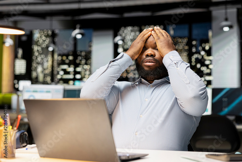 Obraz na płótnie Frustrated man rubs his forehead and feels overburdened, expressing weariness and agitation from intense workload and overtime late at night while feeling anxious and overwhelmed during office work