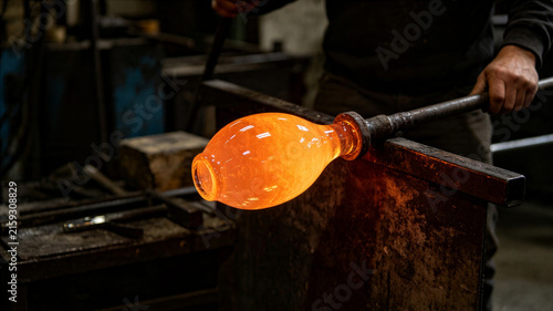 Artisan shaping molten glowing orange glass on blowpipe in workshop