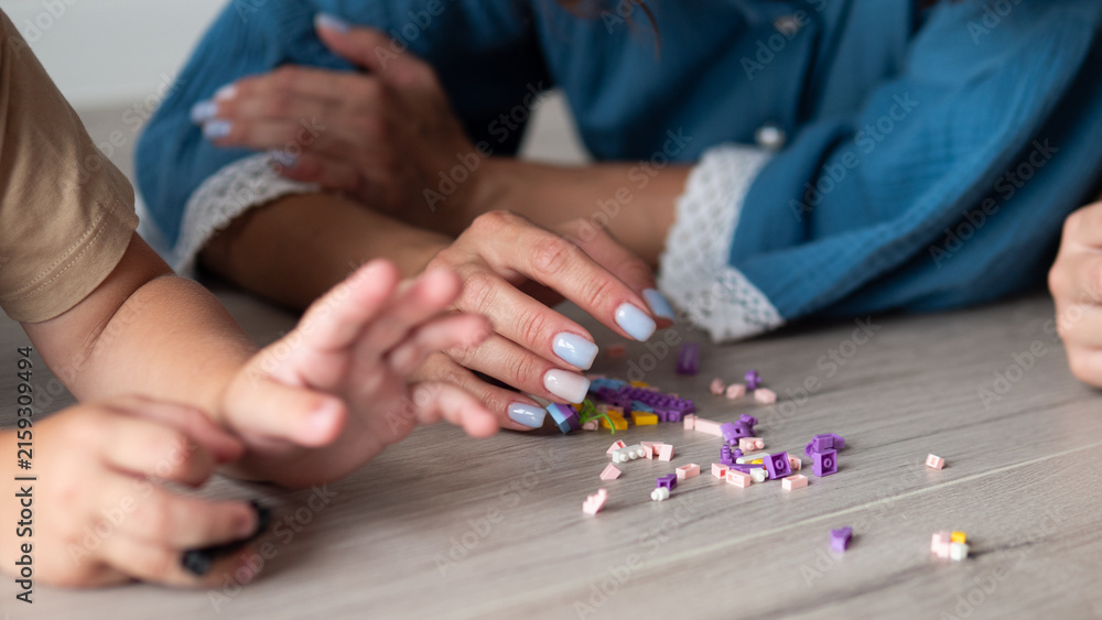 Fototapeta premium Close view of small hands and adult hands reaching toward scattered colorful building pieces on wooden floor, showing cooperative play, home learning, care, creativity, and gentle family connection