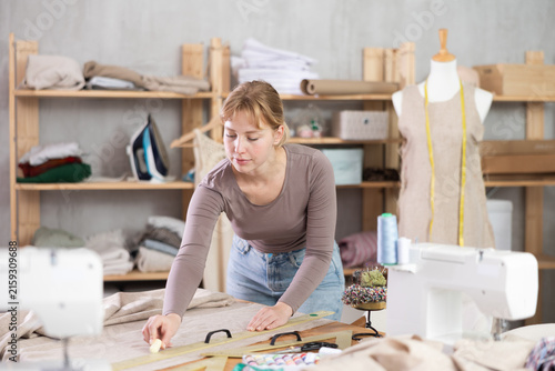 Canvas Print In sewing studio, professional young female dressmaker carefully drawing chalk lines across linen fabric, measuring and marking linen fabric in preparation for cutting