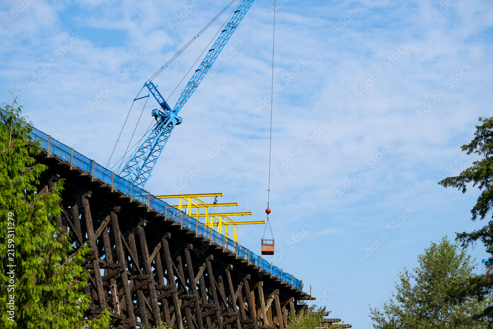 Fototapeta premium Old wooden railroad train trestle construction project, blue crane holding work basket over job site, maintenance on structural framework, sunny summer day, United States