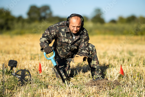 Fotografie A man kneels in a field wearing a camouflage outfit while using a metal detector