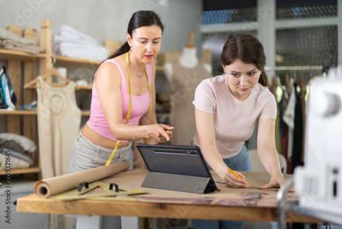 Bild auf Leinwand Two female dressmakers draw outline of future dress on paper while looking at th