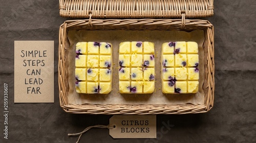 Three fresh lemon blueberry citrus blocks are placed inside a rustic wicker basket on a textured brown fabric background with a motivational quote card and a descriptive paper tag for advertising ads.