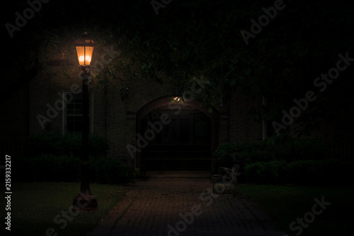 Warm vintage street lamp post illuminating dark cobblestone pathway leading to stone archway at night