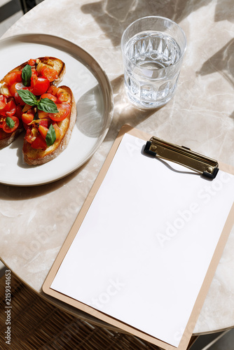 White menu sheet on a clipboard next to tomato toast and water, perfect for cafe menu layouts and restaurant presentation