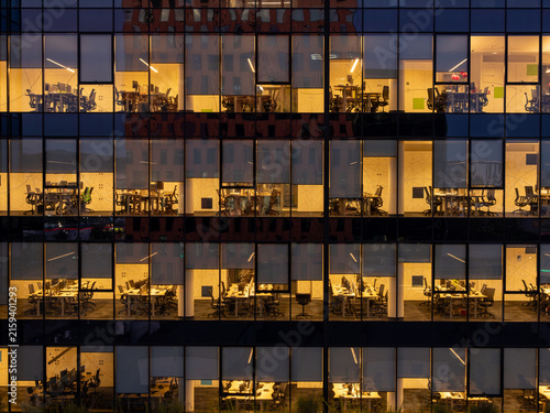 Aerial view of a modern office building facade at night with illuminated windows revealing desks and computer workstations in Gdansk, Poland.