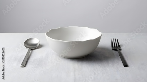 Empty white ceramic bowl with wavy rim, flanked by polished silver spoon and four-tine fork, precisely arranged on a pristine white tablecloth, creating a minimalist and inviting dining setup