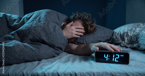 Tired man struggling with insomnia in bed at night reaching to turn off glowing digital alarm clock showing early time