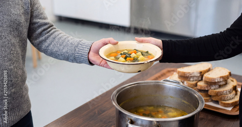 Vegetable soup served in enamel bowl being handed over during a community food charity event in a busy kitchen setting