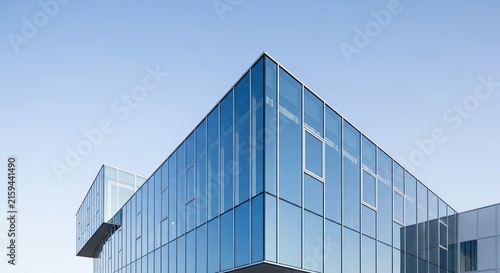 A modern glass building stands against a clear blue sky. The building features large, reflective windows and a sleek, angular design.