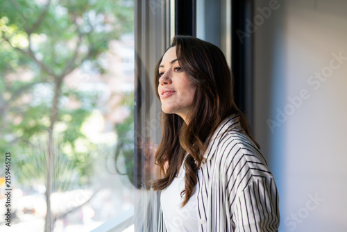 Quadro su tela Young woman standing indoors by a window, looking outside with a pensive and tho
