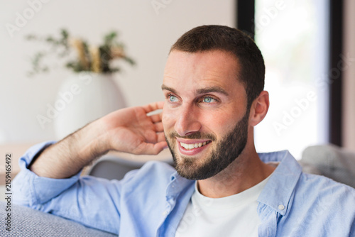 Canvas Print Smiling bearded man sits relaxed on a sofa in a bright living room, wearing a casual blue shirt